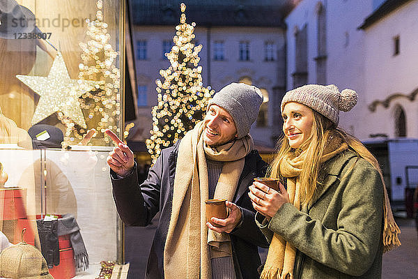 Young couple with cups of mulled wine looking at shop window at Christmas time