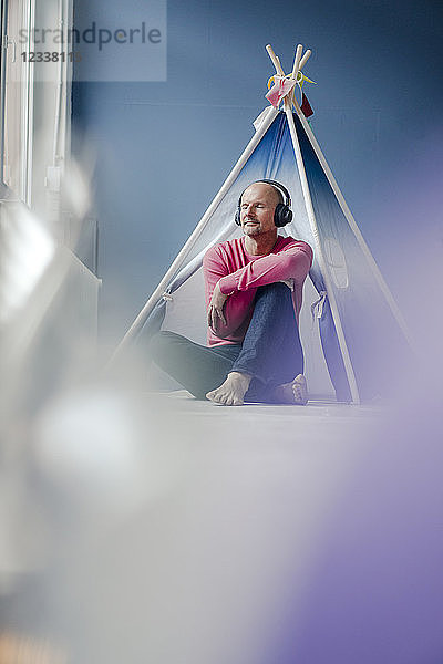 Relaxed mature man wearing headphones sitting at teepee indoors