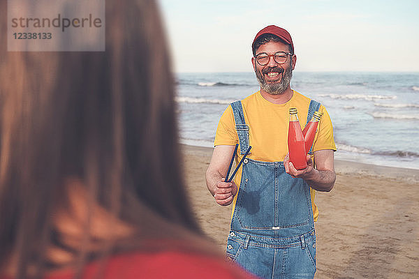 Happy man in dungarees on the beach offering soft drink to woman