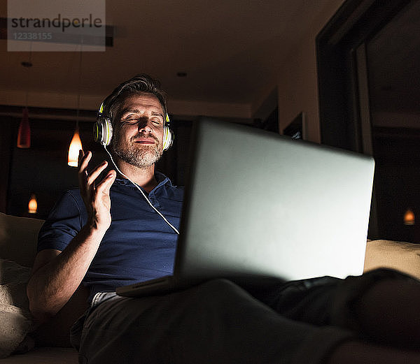 Man sitting on couch at home listening music with headphones and laptop