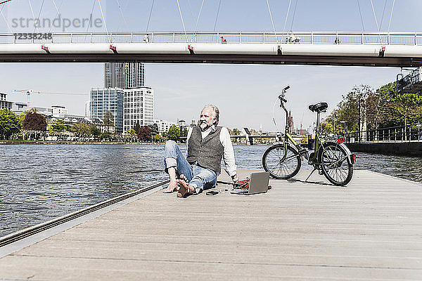Smiling mature man with laptop and bicycle sitting at the riverside in the city