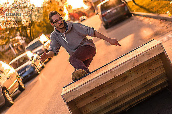 Junger männlicher Skateboarder dreht sich bei Sonnenuntergang auf der Vorstadtstraße und fährt die Rampe hoch