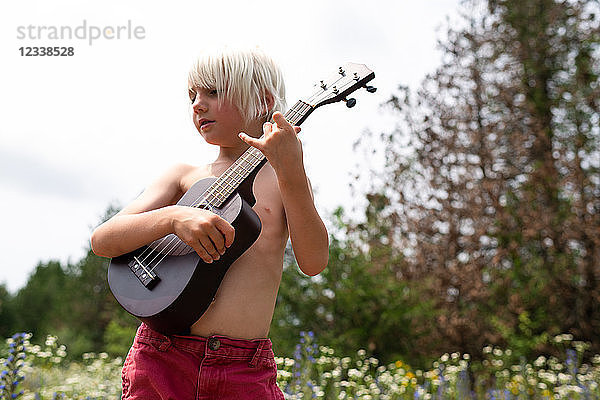 Blondhaariger Junge mit nackter Brust spielt Ukulele auf einer Wildblumenwiese