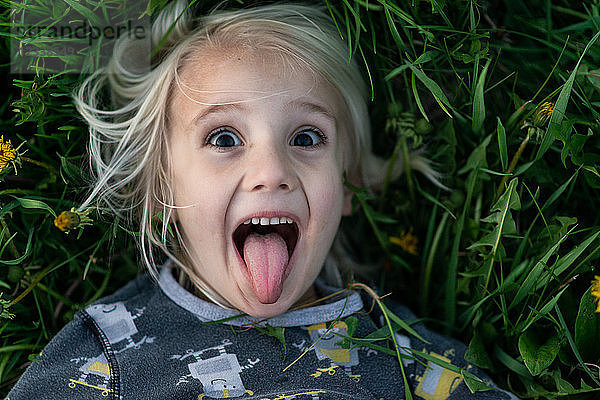 Blondhaariger Junge auf Gras liegend  Zunge herausgestreckt  Porträt