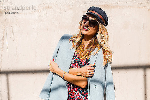 Stilvolle Frau mit Sonnenbrille und Mütze vor der Wand mit verschränkten Armen