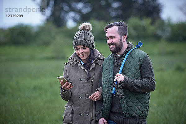 Mann und Frau gehen Arm in Arm über das Feld und schauen auf ein Smartphone
