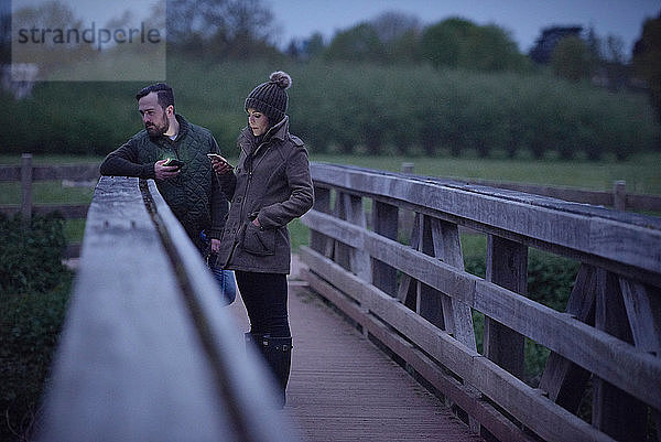 Mann und Frau auf ländlicher Brücke in der Dämmerung beim Blick auf Smartphones