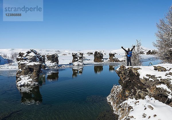 Junger Mann streckt seine Arme in die Luft  Blick auf vulkanische Felsformation im Wasser  Mývatn-See im Winter  Halbinsel Höfði  Island  Europa