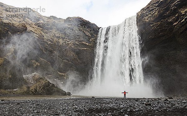 Wanderer mit roter Jacke vor einem hohen Wasserfall Skogafoss  Skogar  Südisland  Island  Europa