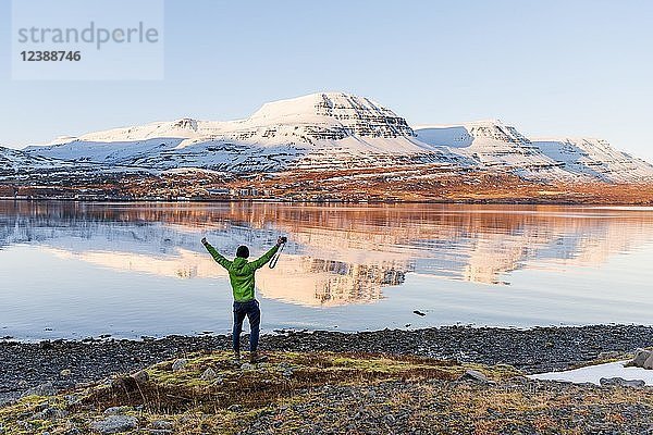 Mann steht am Ufer und streckt die Arme in die Luft  Wasserspiegelung  schneebedeckte Berge in einem Fjord  Reyðarfjörður  Ostisland  Island  Europa