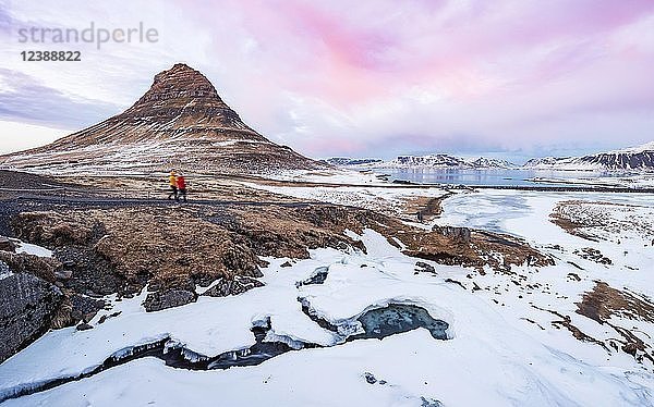 Wanderer  Berg Kirkjufell  Wasserfall Kirkjufellfoss zugefroren  bewölkter Himmel mit Sonnenuntergang  Fjord Grundarfjördur  Westisland  Island  Europa