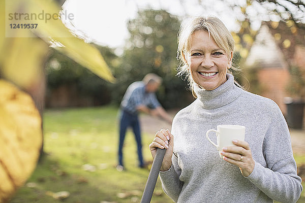 Porträt einer lächelnden  selbstbewussten reifen Frau  die Kaffee trinkt und Herbstblätter im Garten harkt