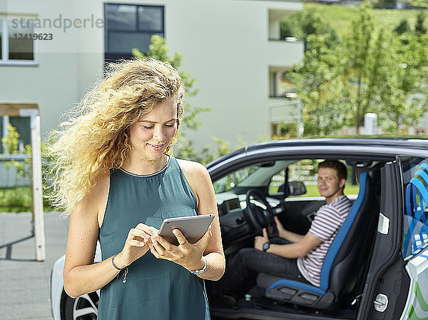 Smiling young woman using tablet with man sitting in electric car in background