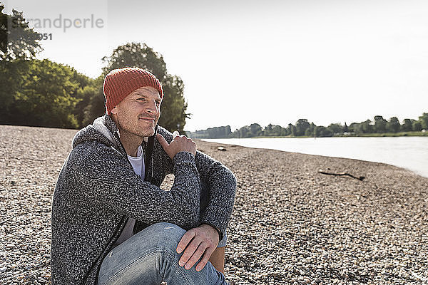 Mature man wearing red beanie sitting at Rhine riverbank