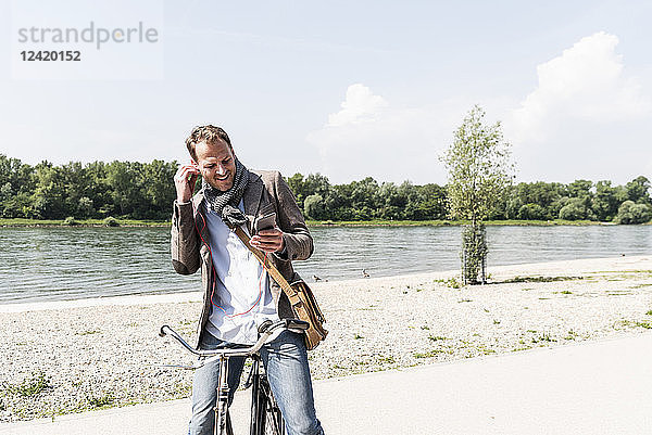 Mature man with bike using smartphone and headphones at Rhine riverbank