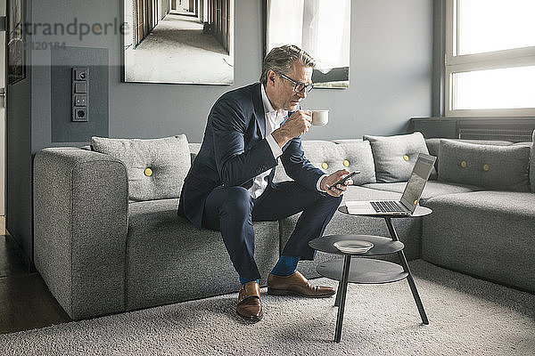 Mature businessman with cup of coffee and laptop using cell phone on couch