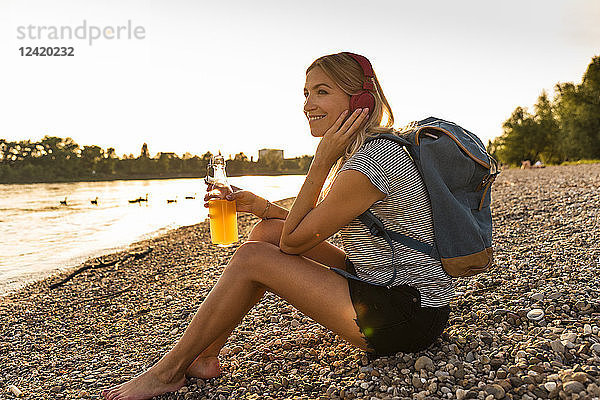 Blond woman with earphone and bottle sitting on riverside in the evening  listening music