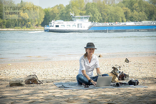 Smiling woman sitting on blanket at a river with dog using laptop