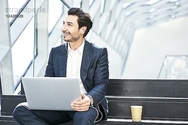 Smiling businessman sitting on stairs wearing earphones and using laptop