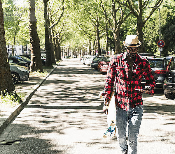 Cool young man with skateboard walking in the city  using smartphone