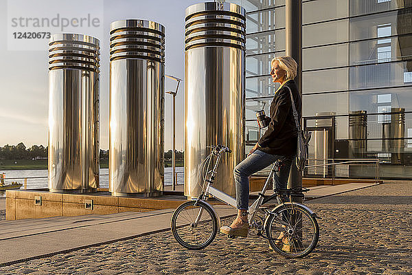 Senior woman with takeaway coffee and city bike having a break at sunset