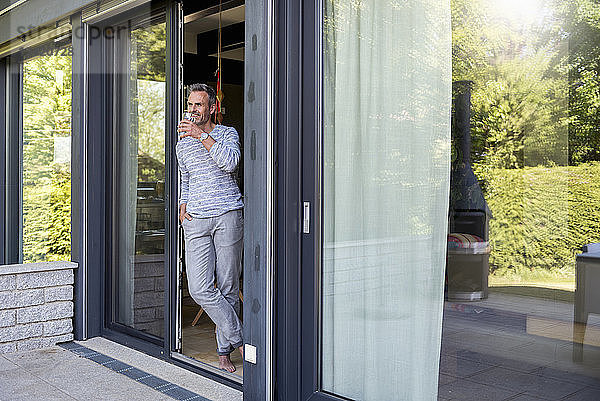 Smiling mature standing at French door at home drinking from glass