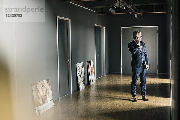 Mature businessman standing on office floor with paintings using cell phone