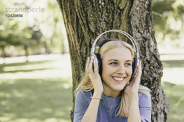 Happy young woman at tree trunk in a park wearing headphones