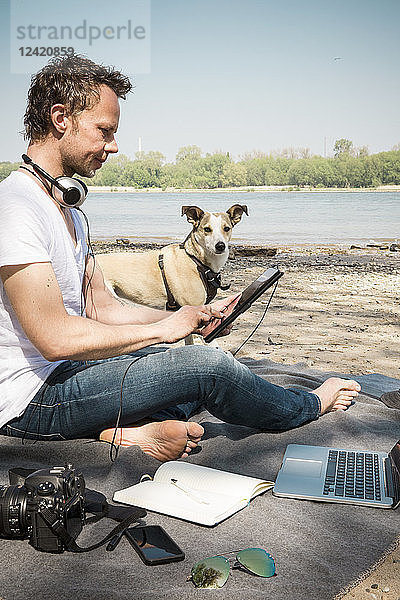 Man with dog sitting on blanket at a river using tablet