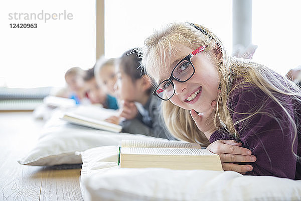 Portrait of smiling schoolgirl lying on the floor with classmates reading book in school break room