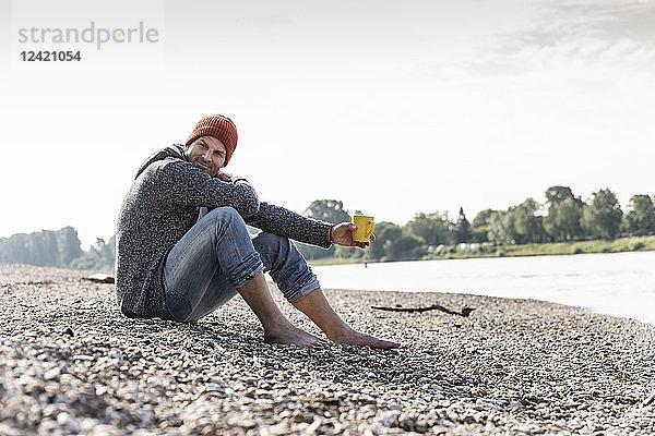 Mature man wearing red beanie and drinking coffee at Rhine riverbank