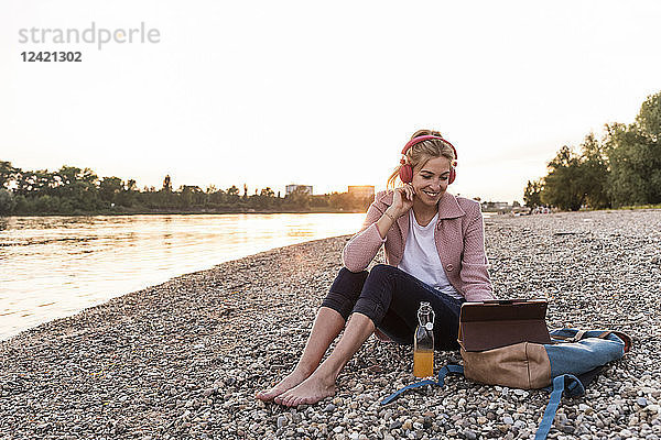 Young blond woman with earphones and tablet on riverside in the evening