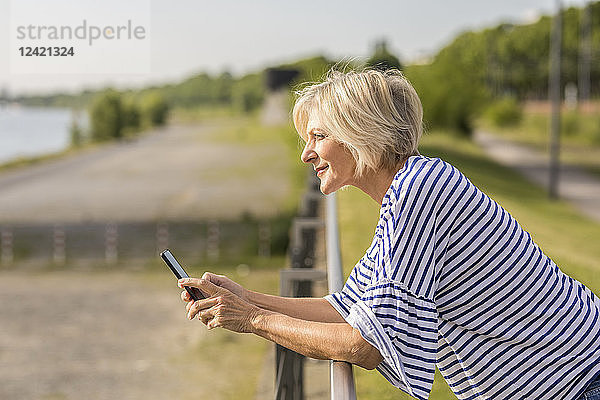 Smiling senior woman with cell phone leaning on railing at riverbank