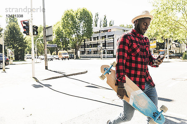 Cool young man with skateboard walking in the city  using smartphone
