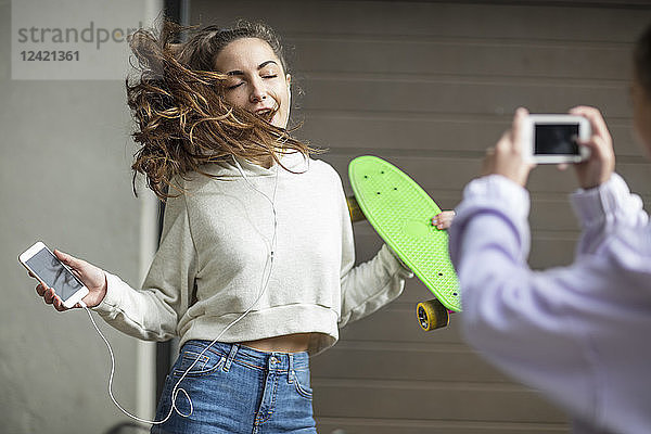 Friend taking picture of carefree teenage girl dancing while holding skateboard and listening to music