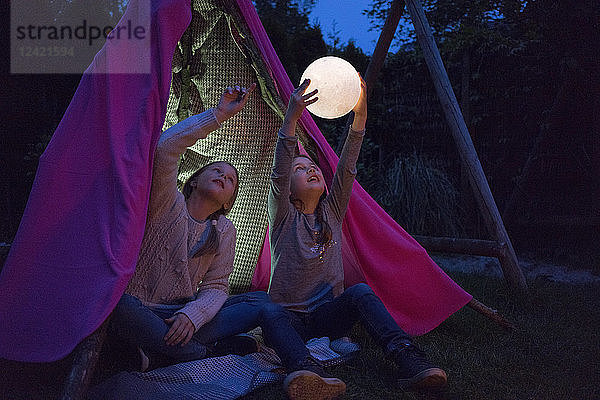 Two girls sitting in tipi  holding lamp as moon