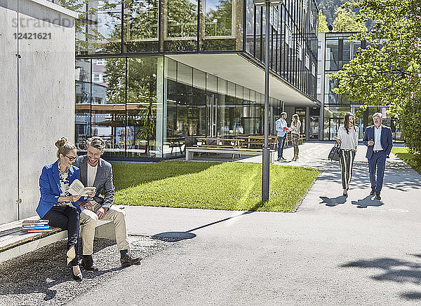 Smiling colleagues with book sitting on bench outside office building