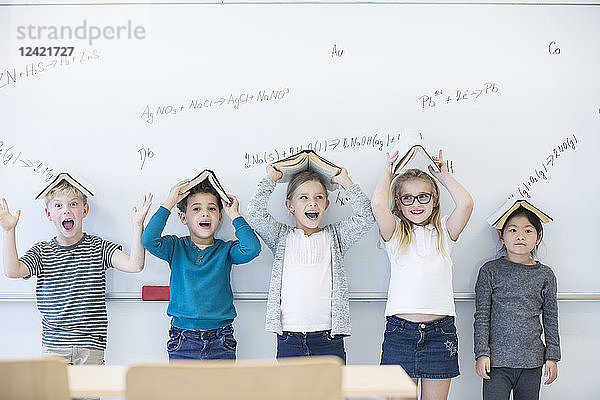 Happy pupils with books above their heads standing at whiteboard with formulas in class