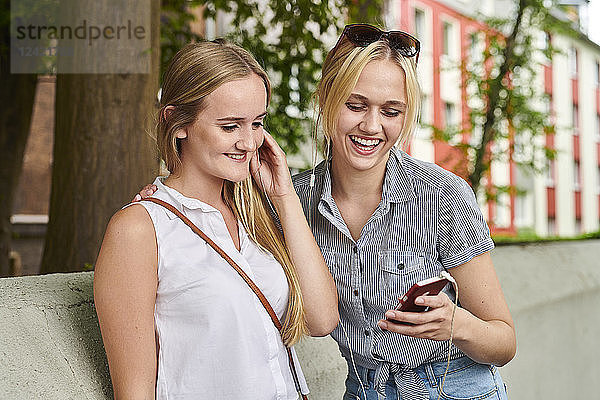 Two happy young women sharing cell phone and earphones outdoors