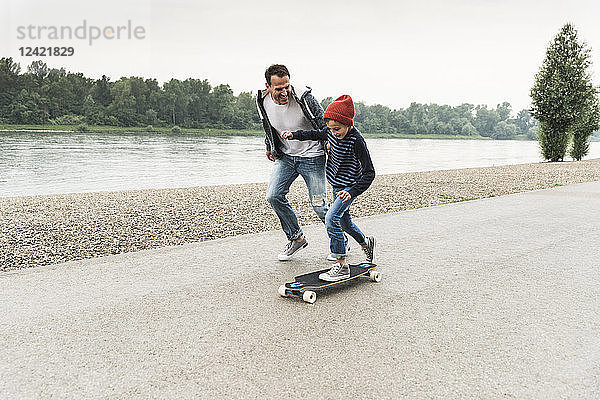Happy father running next to son on skateboard at the riverside