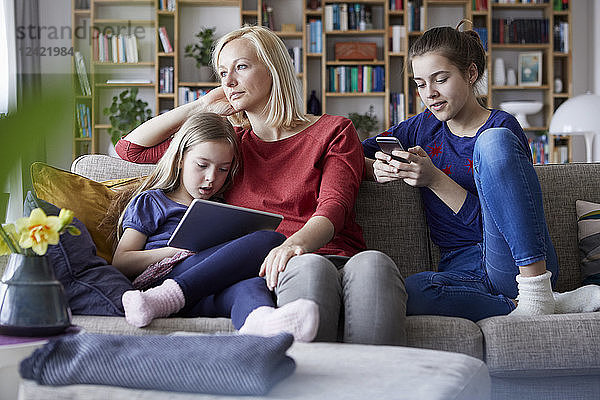 Sad mother sitting on couch with her daughters  playing with mobile devices