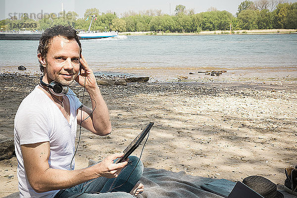 Portrait of smiling man sitting on blanket at a river using tablet