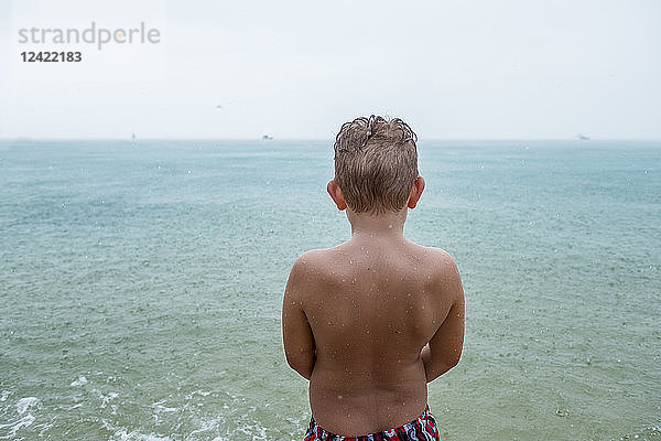 Back view of boy standing in front of the sea on rainy day