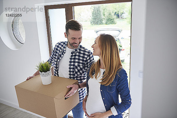 Happy man and pregnant woman moving into new flat carrying cardboard box
