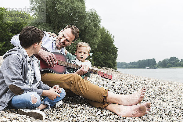 Happy father with two sons sitting at the riverside playing guitar