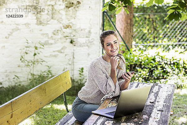 Happy daydreaming woman sitting in the garden with laptop and cell phone