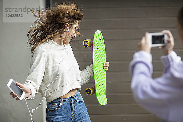 Friend taking picture of carefree teenage girl dancing while holding skateboard and listening to music