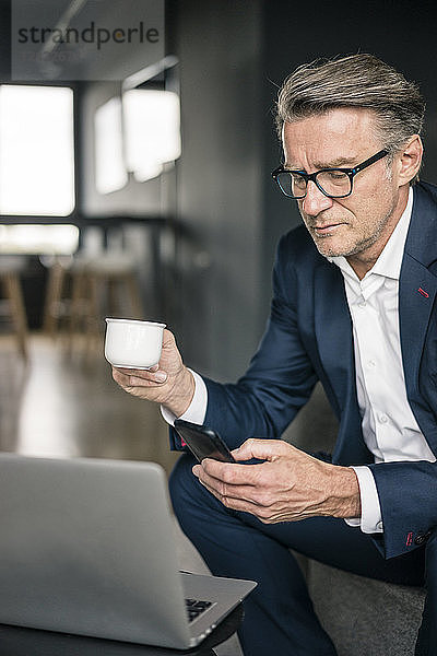 Mature businessman with cup of coffee and laptop using cell phone