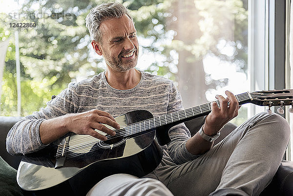 Happy mature man lying on couch at home playing guitar