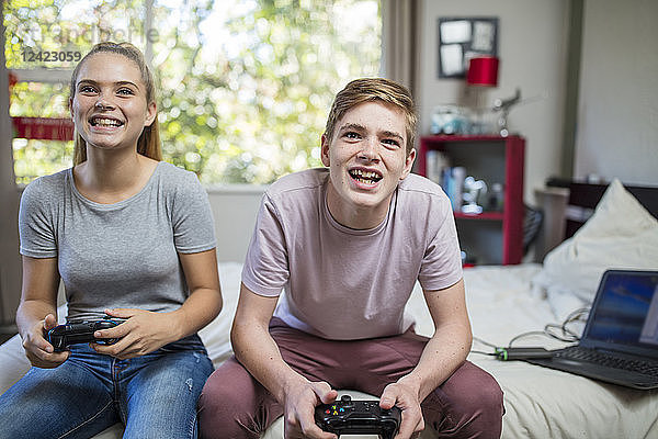 Happy teenage girl and boy sitting on bed playing video game